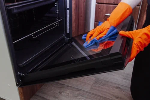 Cleaner in orange gloves wiping the inside of an open oven door during a kitchen deep clean